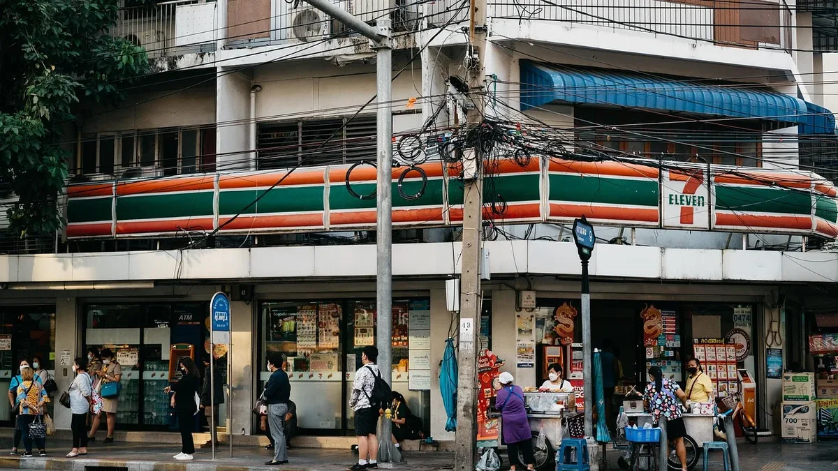 A 7-Eleven convenience store with a balcony and a street in front of it, featuring people walking and standing outside.