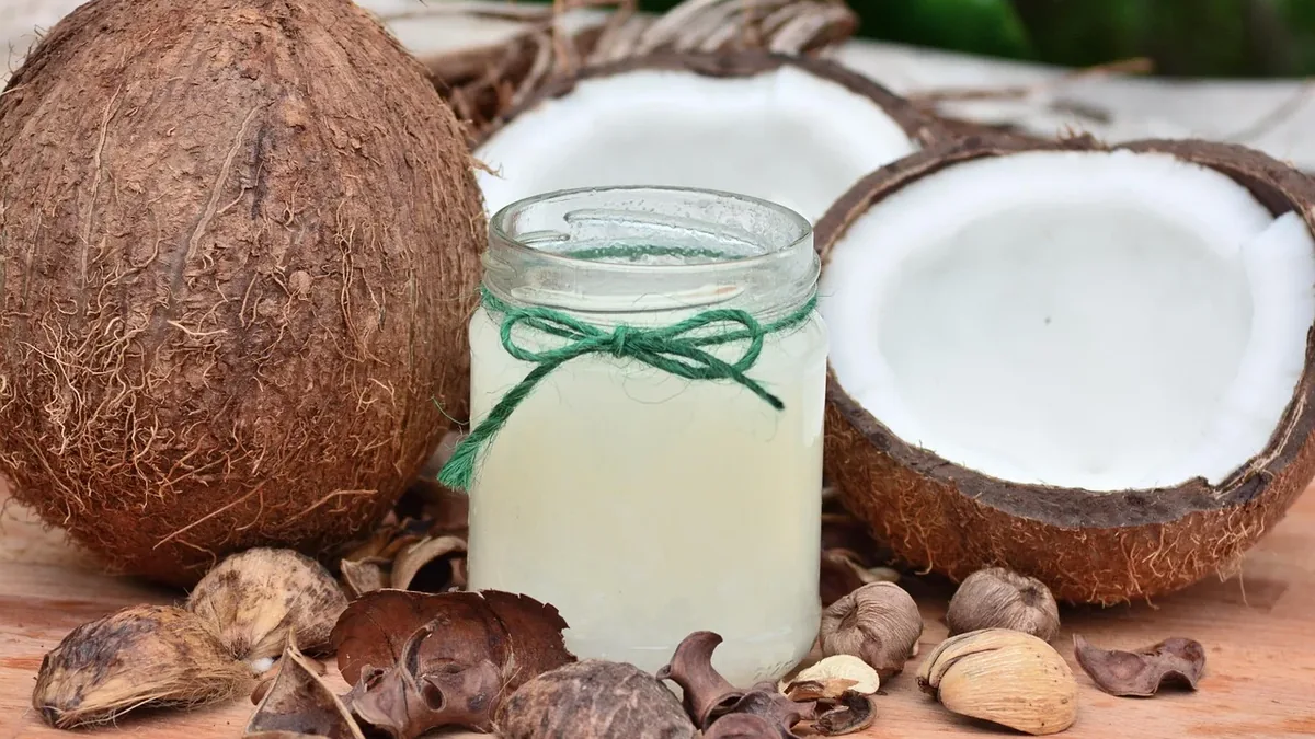 A glass jar filled with coconut oil, surrounded by a coconut shell, a halved coconut, and various coconut husk pieces on a wooden surface.