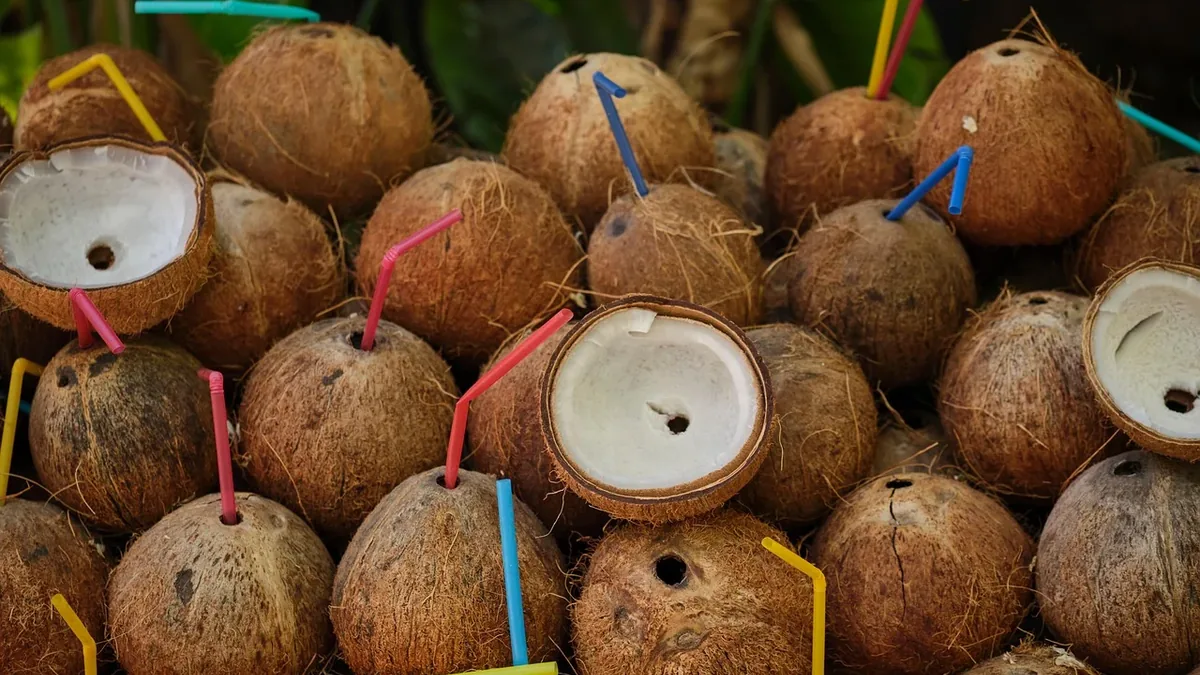 A pile of coconuts with straws inserted into them, set against a backdrop of green leaves.