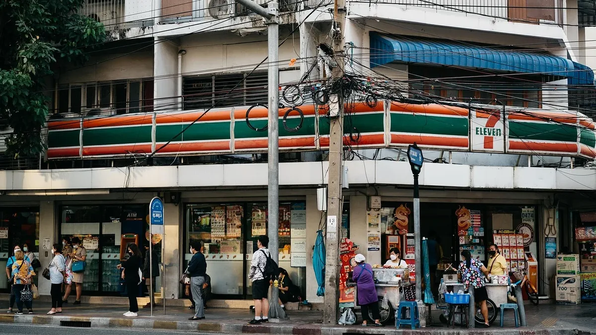 A 7-Eleven convenience store with a balcony and a street in front of it, featuring people walking and standing outside.