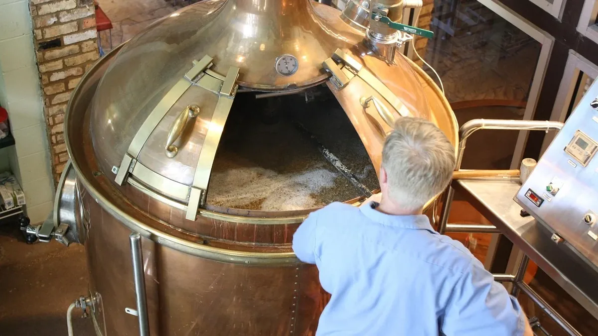 A man standing in front of a large copper-colored vat with a hinged lid, which appears to be used for brewing beer.