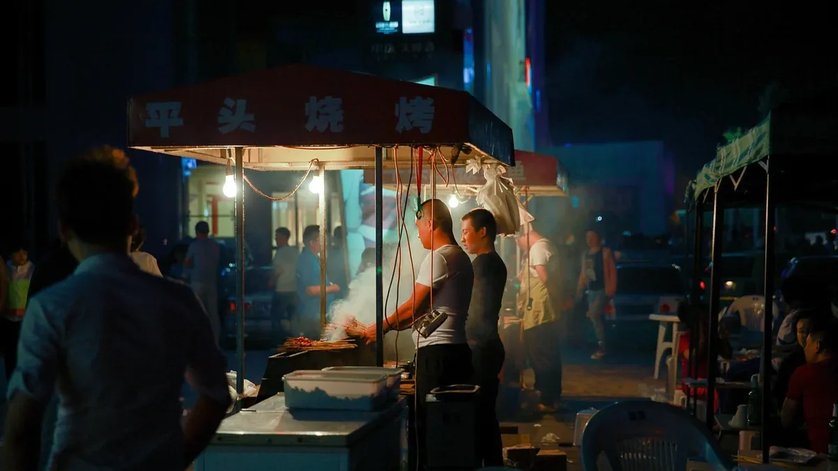 A nighttime street food stall with a red canopy, where a man is cooking on a grill, and a crowd of people are gathered around the stall.