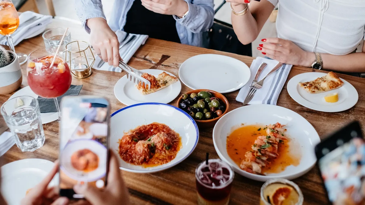 A table with a variety of food and drinks, with people sitting around it, taking pictures of their meals with their phones.
