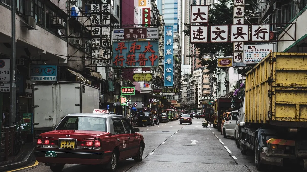 A busy street in Hong Kong, lined with tall buildings and featuring a red taxi, a yellow truck, and a white van, with a multitude of signs a