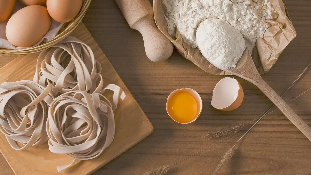 A wooden table with a bowl of eggs, a cutting board with noodles, a rolling pin, a bag of flour, a wooden spoon, an eggshell, and wheat stal