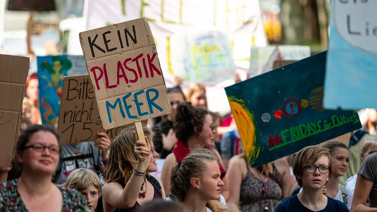 A crowd of people is gathered, with some holding signs and banners, and one person prominently holding a sign that reads "KEIN PLASTIK MEER"