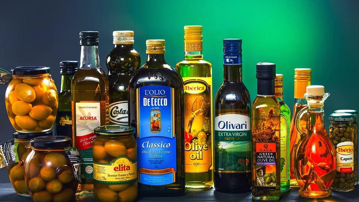 A collection of various bottles of olive oil and jars of olives on a table against a green background.