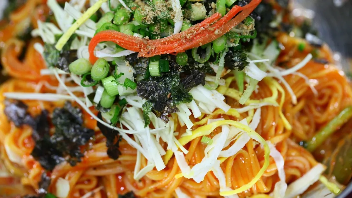 A close-up of a bowl of noodles topped with a variety of vegetables, including shredded white and yellow cabbage, green onions, and what app