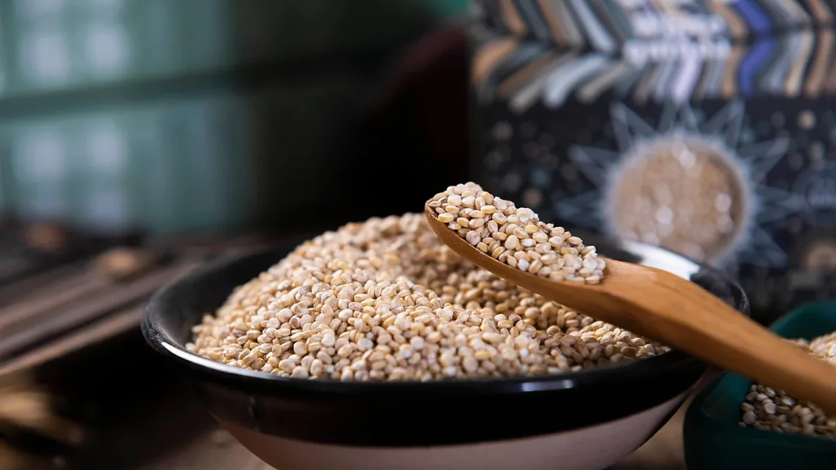 A bowl of sesame seeds with a wooden spoon resting on top, set against a blurred background that appears to be a room with a table and a dec