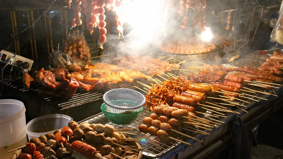 A street food stall with various skewers of meat and vegetables being grilled over an open flame.