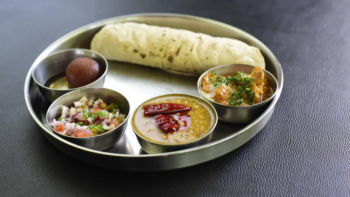 A stainless steel plate containing a variety of Indian dishes, including a rolled flatbread, a small bowl of rice, a small bowl of soup, a s